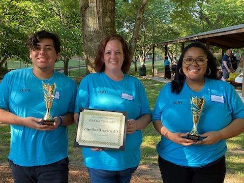 Three individuals in blue "TRIO" shirts holding awards.